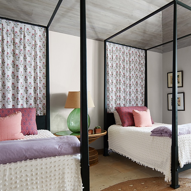 Two black canopy twin beds fit nicely in this off-white painted bedroom with a vaulted shiplap ceiling, colorful pastel and white décor, and a door open to the outside. Two black canopy twin beds fit nicely in this off-white painted bedroom with a vaulted shiplap ceiling, colorful pastel and white décor, and a door open to the outside.