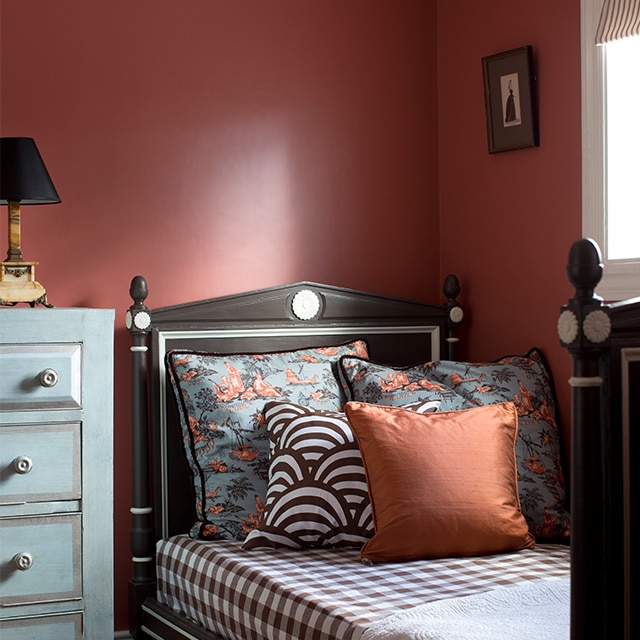 An inviting corner bedroom space with warm red painted walls, a blue dresser and a single bed with eclectic print bedding underneath a white trimmed window. An inviting corner bedroom space with warm red painted walls, a blue dresser and a single bed with eclectic print bedding underneath a white trimmed window.