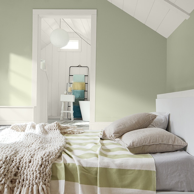 A sunlit sage green painted bedroom features a white ceiling and trim, a white bed with a green and white striped blanket, and an open door into a white painted en suite bathroom. A sunlit sage green painted bedroom features a white ceiling and trim, a white bed with a green and white striped blanket, and an open door into a white painted en suite bathroom.