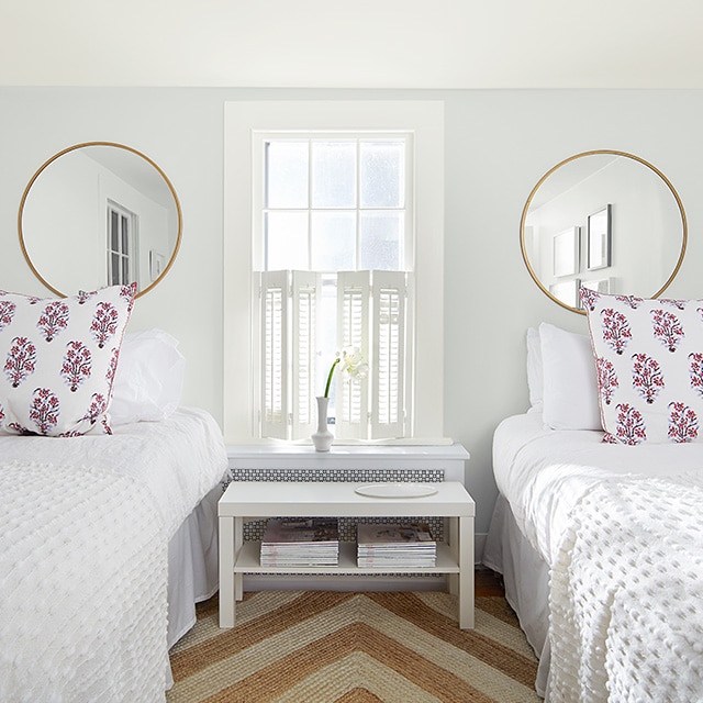 A white painted bedroom with two circular wall mirrors, twin beds with white bedding, a white shuttered window and striped jute rug. A white painted bedroom with two circular wall mirrors, twin beds with white bedding, a white shuttered window and striped jute rug.