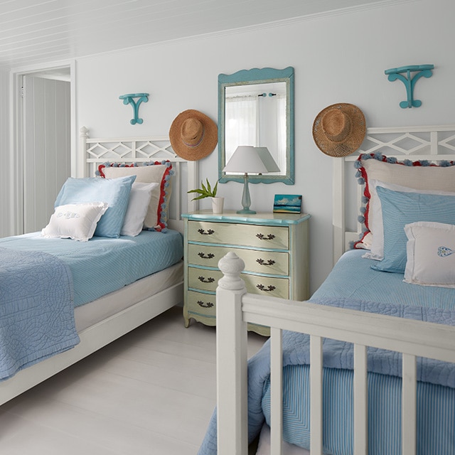 A cheerful, white-painted bedroom features two twin beds with white bed frames and blue bedding, flanking a pale yellow-green dresser with blue trim and a blue framed mirror. A cheerful, white-painted bedroom features two twin beds with white bed frames and blue bedding, flanking a pale yellow-green dresser with blue trim and a blue framed mirror.
