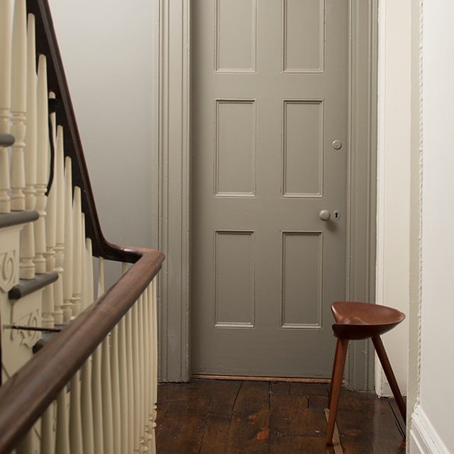 A hallway painted in neutrals with a wooden floor, a banister with off-white spindles, a gray-painted door, and wooden stool. A hallway painted in neutrals with a wooden floor, a banister with off-white spindles, a gray-painted door, and wooden stool.