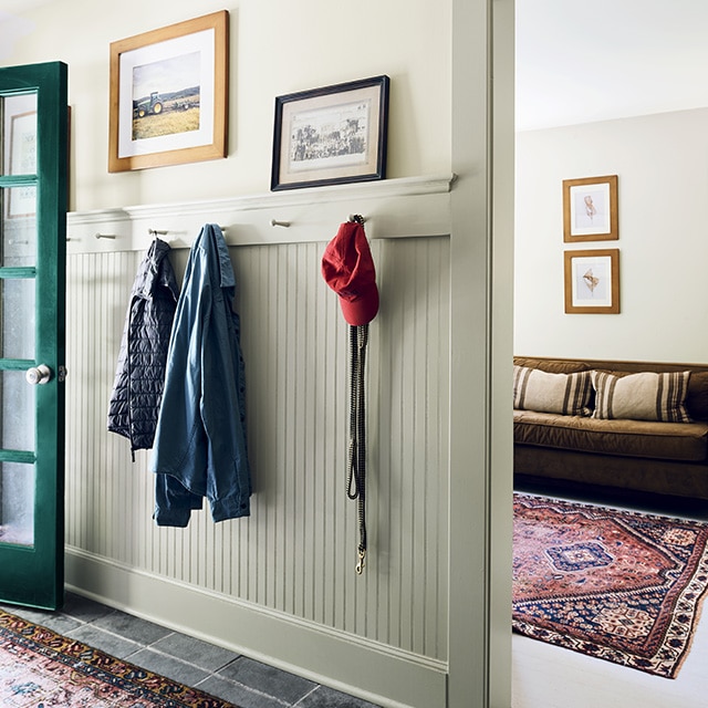 A sunlit entry hallway with a dark green front door, off-white walls, pale green painted wainscoting and trim, and a side room in neutral tones seen through an open doorway. A sunlit entry hallway with a dark green front door, off-white walls, pale green painted wainscoting and trim, and a side room in neutral tones seen through an open doorway.