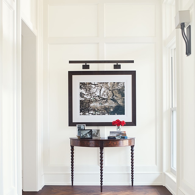 A bright, white-painted hallway with elegant board and batten walls, a piece of framed art, a table, tan striped rug, and dark wood floor. A bright, white-painted hallway with elegant board and batten walls, a piece of framed art, a table, tan striped rug, and dark wood floor.