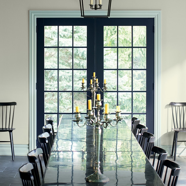 Beautiful dark navy blue-painted French doors with blue-gray trim stand out in this sunlit cool gray-painted dining room with a long black table and chairs. Beautiful dark navy blue-painted French doors with blue-gray trim stand out in this sunlit cool gray-painted dining room with a long black table and chairs.