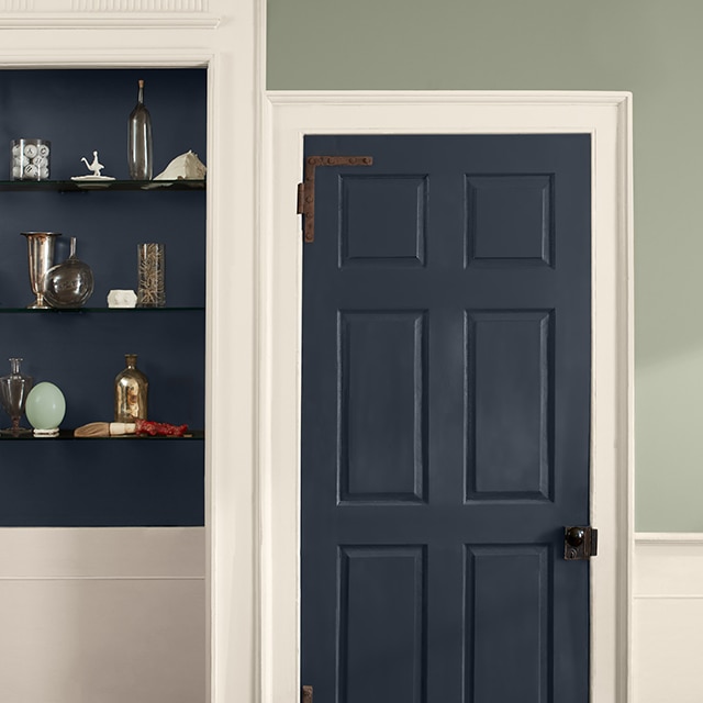 A two-tone wall painted light-sage green on the upper wall and off-white on the lower wall, with off-white trim and a dark blue door and inset shelves holding glass vases and trinkets. A two-tone wall painted light-sage green on the upper wall and off-white on the lower wall, with off-white trim and a dark blue door and inset shelves holding glass vases and trinkets.