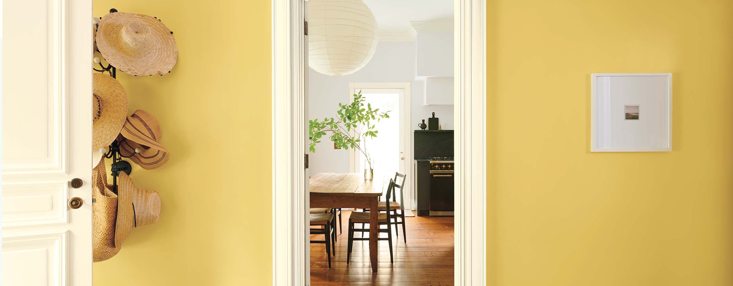A pretty golden yellow-painted entryway with a mounted hat rack, and a white open door and trim, looking into a light gray-painted kitchen. A pretty golden yellow-painted entryway with a mounted hat rack, and a white open door and trim, looking into a light gray-painted kitchen.