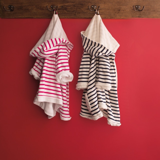 Mur rouge avec portemanteau mural en bois où sont accrochés deux petits peignoirs rayés, l’un en rose et l’autre en noir. Mur rouge avec portemanteau mural en bois où sont accrochés deux petits peignoirs rayés, l’un en rose et l’autre en noir.