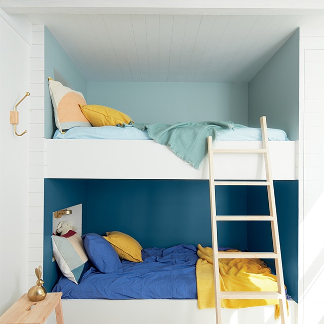 Chambre blanche avec deux lits encastrés, l’un au-dessus de l’autre, chacun peint dans sa propre nuance de bleu avec literie assortie, coussins jaunes, tabouret-escabeau en bois et échelle. Chambre blanche avec deux lits encastrés, l’un au-dessus de l’autre, chacun peint dans sa propre nuance de bleu avec literie assortie, coussins jaunes, tabouret-escabeau en bois et échelle.