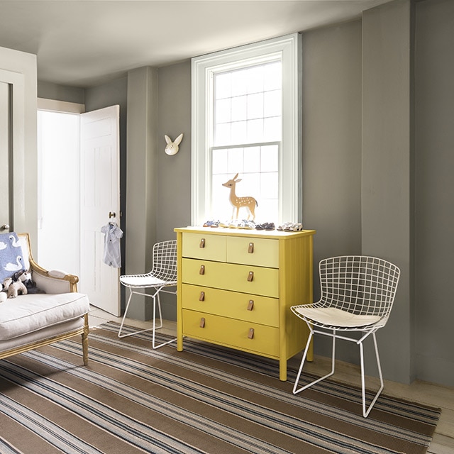 Salle aux murs gris, moulures et portes blanches, commode en bois jaune ombré flanquée de deux chaises en métal blanc et sofa beige avec animaux en peluche. Salle aux murs gris, moulures et portes blanches, commode en bois jaune ombré flanquée de deux chaises en métal blanc et sofa beige avec animaux en peluche.
