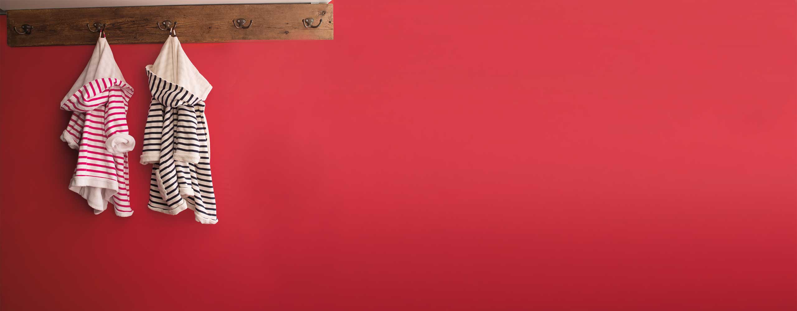 Mur rouge avec portemanteau mural en bois où sont accrochés deux petits peignoirs rayés, l’un en rose et l’autre en noir. Mur rouge avec portemanteau mural en bois où sont accrochés deux petits peignoirs rayés, l’un en rose et l’autre en noir.