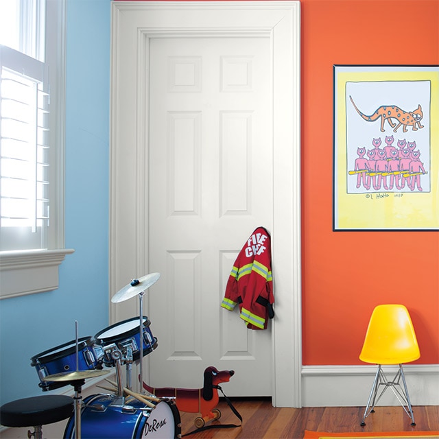 Salle de jeux à l’agencement complémentaire – un mur orange et un mur bleu – porte et moulures blanches, batterie, dessin encadré et chaise jaune. Salle de jeux à l’agencement complémentaire – un mur orange et un mur bleu – porte et moulures blanches, batterie, dessin encadré et chaise jaune.