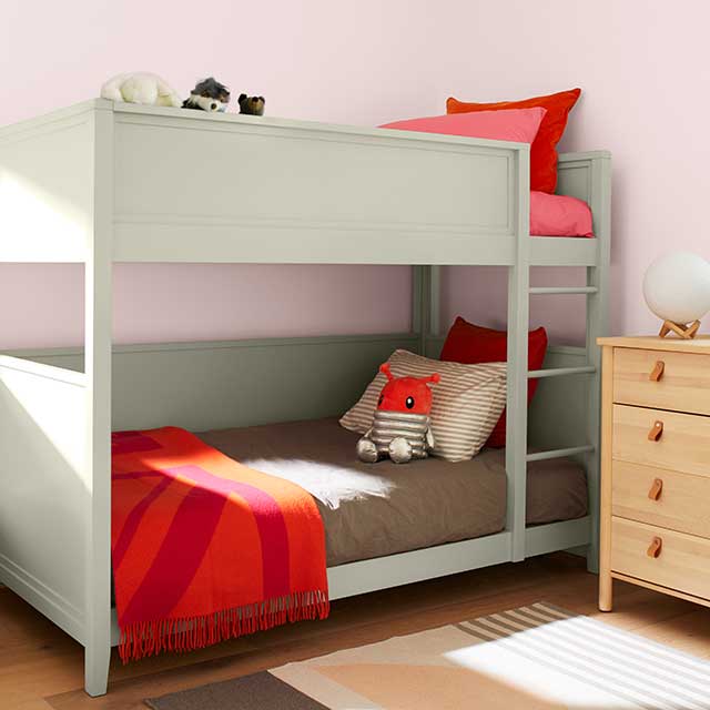 Chambre d’enfant rose pâle avec lit superposé jade aux articles de literie gris et rouges, commode en bois et tapis à rayures grises. Chambre d’enfant rose pâle avec lit superposé jade aux articles de literie gris et rouges, commode en bois et tapis à rayures grises.