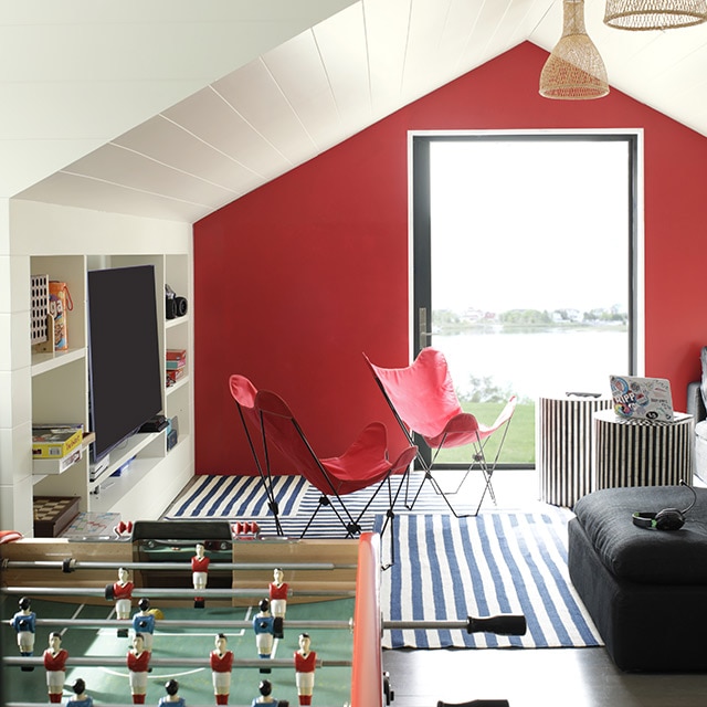 Salle de jeux blanche avec étagères encastrées dont une pour le téléviseur, plafond voûté en planches à feuillure, mur d’accent rouge avec porte vitrée, table de billard et jeu de soccer sur table, sofa noir et fauteuils rouges. Salle de jeux blanche avec étagères encastrées dont une pour le téléviseur, plafond voûté en planches à feuillure, mur d’accent rouge avec porte vitrée, table de billard et jeu de soccer sur table, sofa noir et fauteuils rouges.