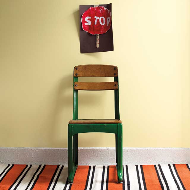 Chaise d’école verte avec siège et dossier en bois devant un mur jaune pâle avec dessin d’un panneau d’arrêt accroché au mur et petit tapis aux rayures orange, blanc et noir. Chaise d’école verte avec siège et dossier en bois devant un mur jaune pâle avec dessin d’un panneau d’arrêt accroché au mur et petit tapis aux rayures orange, blanc et noir.
