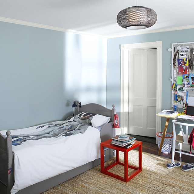 Chambre d’enfant aux murs bleu pâle avec moulures, plafond et porte blancs ainsi qu’un lit gris à la literie d’astronaute, une table rouge et un bureau et une chaise sous un babillard. Chambre d’enfant aux murs bleu pâle avec moulures, plafond et porte blancs ainsi qu’un lit gris à la literie d’astronaute, une table rouge et un bureau et une chaise sous un babillard.