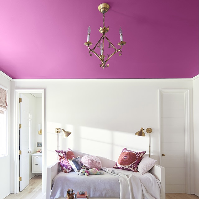 Un plafond rose foncé ajoute un joyeux éclat de couleur dans cette chambre blanche aux coussins multicolores et à la literie blanche sur une base de lit blanche entre deux portes. Un plafond rose foncé ajoute un joyeux éclat de couleur dans cette chambre blanche aux coussins multicolores et à la literie blanche sur une base de lit blanche entre deux portes.