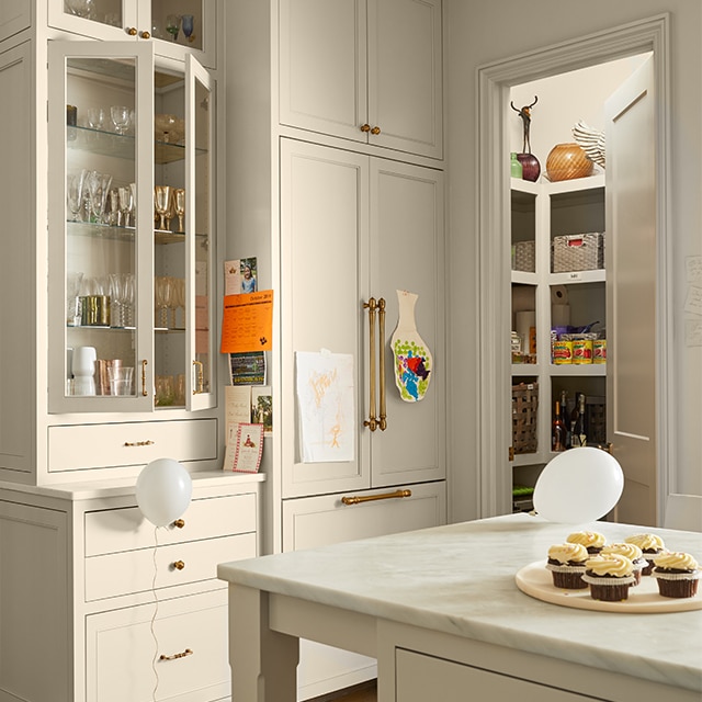 A bright, beige-painted kitchen with floor-to-ceiling and glass cabinets, an island with a marble countertop, an open pantry door, and white balloons on the floor and island. A bright, beige-painted kitchen with floor-to-ceiling and glass cabinets, an island with a marble countertop, an open pantry door, and white balloons on the floor and island.