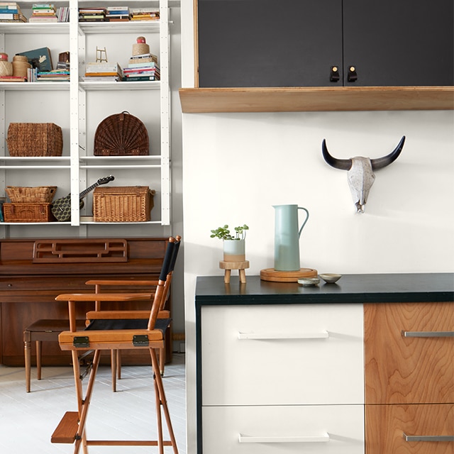 Bright, white-painted loft space with black-painted upper cabinets, a two-tone white and wood lower cabinet, a director’s chair, and tall white shelves in a back room over a piano. Bright, white-painted loft space with black-painted upper cabinets, a two-tone white and wood lower cabinet, a director’s chair, and tall white shelves in a back room over a piano.