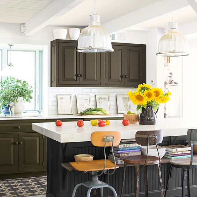 An airy industrial style kitchen taps into deep colors, from a black-based center island with a white countertop to deep, green-brown painted cabinetry. An airy industrial style kitchen taps into deep colors, from a black-based center island with a white countertop to deep, green-brown painted cabinetry.