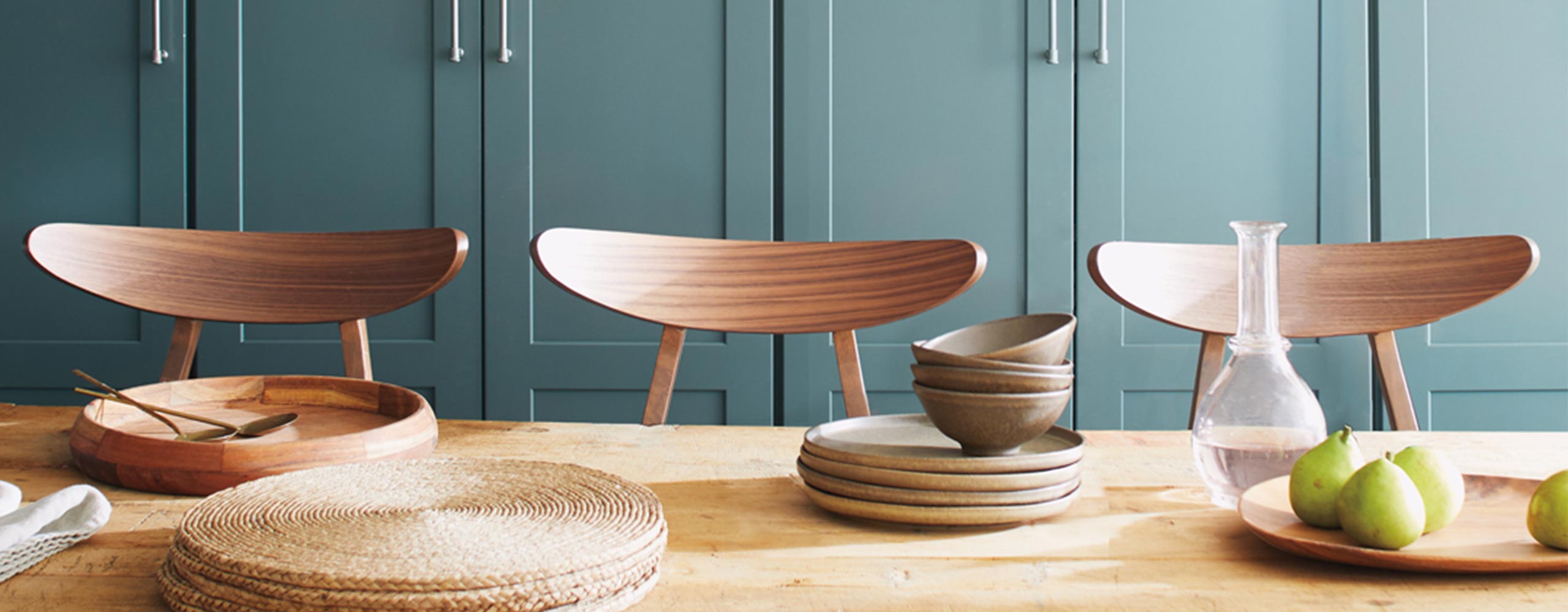 A wall of beautiful light teal-painted kitchen cabinets behind a wooden tabletop with wicker placemats, wooden tableware, and three wood chairs. A wall of beautiful light teal-painted kitchen cabinets behind a wooden tabletop with wicker placemats, wooden tableware, and three wood chairs.