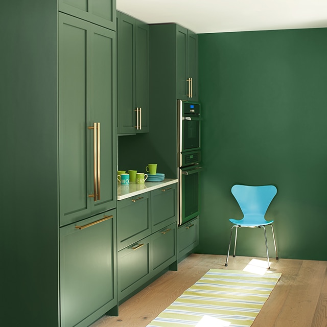 A sleek kitchen space with uniformly green painted built-in cabinets and wall, a white ceiling, modern turquoise chair, and a yellow-striped rug on a blonde wood floor. A sleek kitchen space with uniformly green painted built-in cabinets and wall, a white ceiling, modern turquoise chair, and a yellow-striped rug on a blonde wood floor.