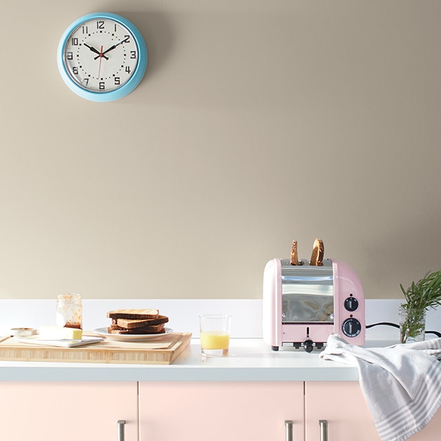 A kitchen with light pink painted cabinets, a pink toaster, cutting board, dishes, and tan painted wall with blue clock. A kitchen with light pink painted cabinets, a pink toaster, cutting board, dishes, and tan painted wall with blue clock.