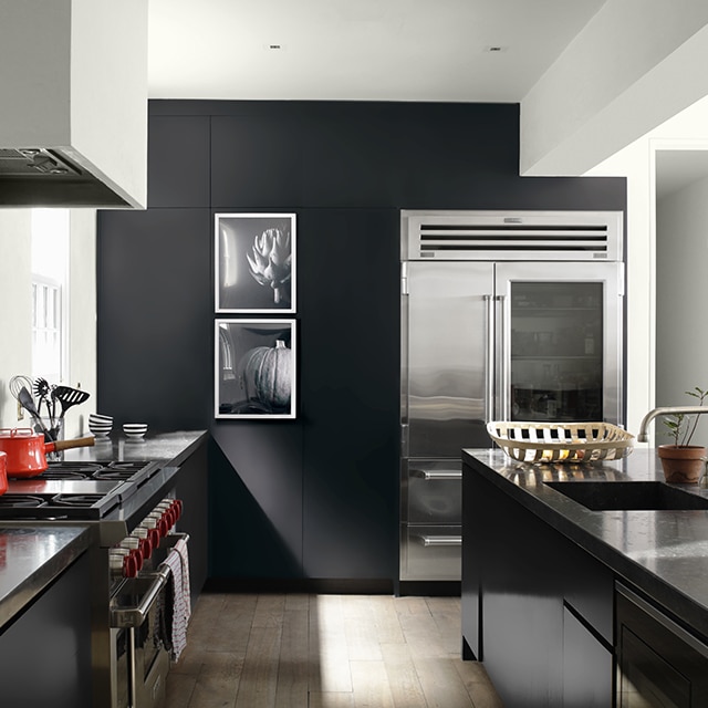 A sleek, contemporary kitchen with white-painted walls and ceiling, a black accent wall, painted black cabinets, and a large island, open to a white hallway. A sleek, contemporary kitchen with white-painted walls and ceiling, a black accent wall, painted black cabinets, and a large island, open to a white hallway.