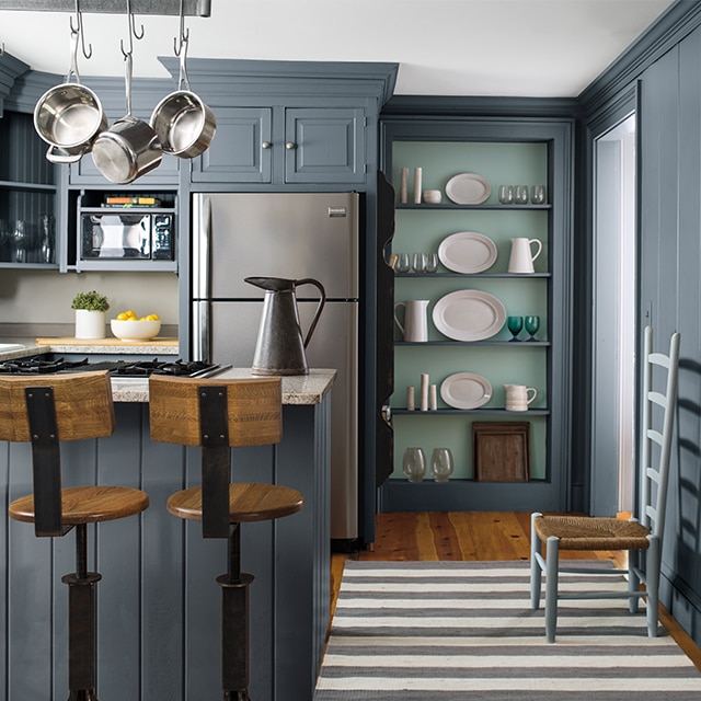 A dark gray painted kitchen featuring beautiful built-in kitchen shelving with a light blue back wall and gray shelves holding white ceramic tableware and glassware, next to a stainless-steel refrigerator. A dark gray painted kitchen featuring beautiful built-in kitchen shelving with a light blue back wall and gray shelves holding white ceramic tableware and glassware, next to a stainless-steel refrigerator.