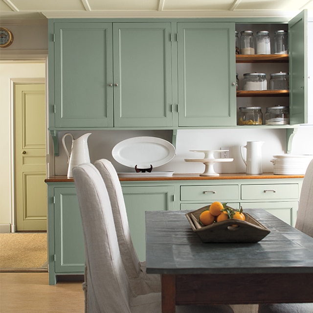 Light green-painted cabinets and white dinnerware add a soft glow as a backdrop to a dining table and white slipcovered chairs. Light green-painted cabinets and white dinnerware add a soft glow as a backdrop to a dining table and white slipcovered chairs.