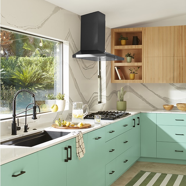 An airy white kitchen with lower cabinets painted in a light mint green, a large picture window over a black sink, white countertops, and upper cabinets with open shelving in light wood. An airy white kitchen with lower cabinets painted in a light mint green, a large picture window over a black sink, white countertops, and upper cabinets with open shelving in light wood.