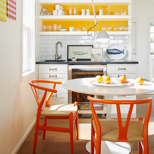 A white-painted kitchen with white cabinets and open shelving, a yellow accent wall, a white table and orange chairs. A white-painted kitchen with white cabinets and open shelving, a yellow accent wall, a white table and orange chairs.