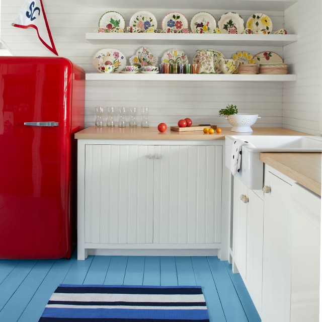 A white-painted kitchen with a light blue-painted floor, red refrigerator and white floating shelves. A white-painted kitchen with a light blue-painted floor, red refrigerator and white floating shelves.
