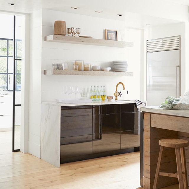 A pleasant, white-painted kitchen with black cabinets, marble countertops, open wood shelving holding tableware, and a wood-based island and stool. A pleasant, white-painted kitchen with black cabinets, marble countertops, open wood shelving holding tableware, and a wood-based island and stool.
