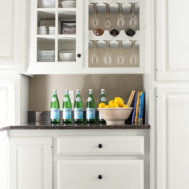 White-painted kitchen cabinets, a built-in wine rack and drawers, with a black countertop displaying water bottles and a bowl of lemons. White-painted kitchen cabinets, a built-in wine rack and drawers, with a black countertop displaying water bottles and a bowl of lemons.