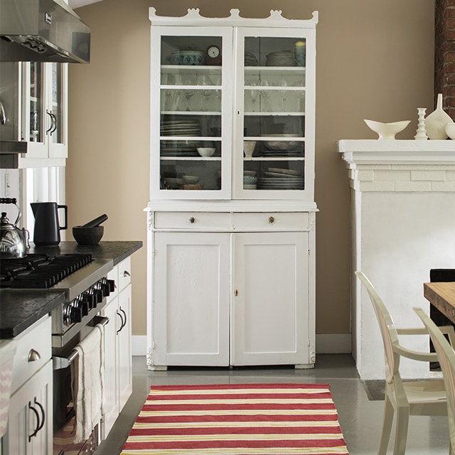 A greige-painted kitchen wall with a tall white hutch, white fireplace, white kitchen cabinets, and a red and white striped rug. A greige-painted kitchen wall with a tall white hutch, white fireplace, white kitchen cabinets, and a red and white striped rug.