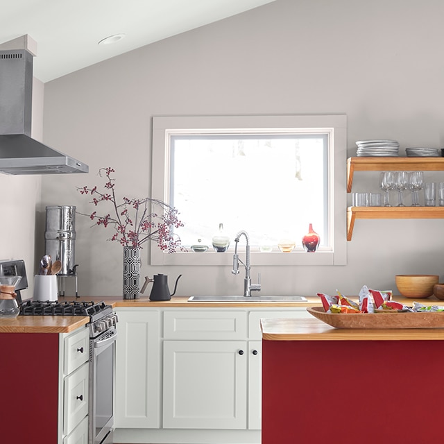 A cheery small kitchen with lavender-gray walls, red-painted cabinet siding, white-painted lower cabinets, wooden floors and a sunny bright window above the sink. A cheery small kitchen with lavender-gray walls, red-painted cabinet siding, white-painted lower cabinets, wooden floors and a sunny bright window above the sink.