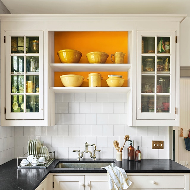 A white painted kitchen with black countertops and an orange accent wall behind open shelving. A white painted kitchen with black countertops and an orange accent wall behind open shelving.