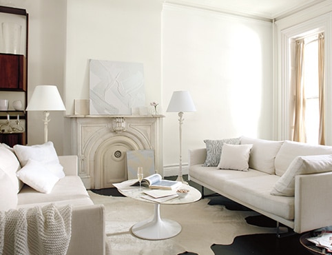 Neutral living room with white walls and an off-white wall mantel, and a white circle coffee table in between two cream couches. Neutral living room with white walls and an off-white wall mantel, and a white circle coffee table in between two cream couches.