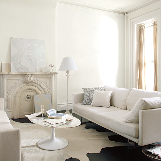 Neutral living room with white walls and an off white wall mantel, and a white circle coffee table in between two cream couches. Neutral living room with white walls and an off white wall mantel, and a white circle coffee table in between two cream couches.