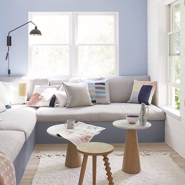 A bright, cheerful periwinkle painted living room with a white side wall and trim, a large banquette sectional with a gray base, and three small tables. A bright, cheerful periwinkle painted living room with a white side wall and trim, a large banquette sectional with a gray base, and three small tables.