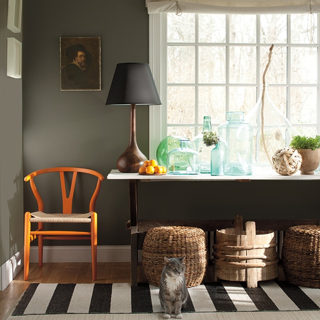 A dark green painted living room corner with white trim, an orange chair, baskets, and a table with bottles and lamp, in front of a picture window. A dark green painted living room corner with white trim, an orange chair, baskets, and a table with bottles and lamp, in front of a picture window.