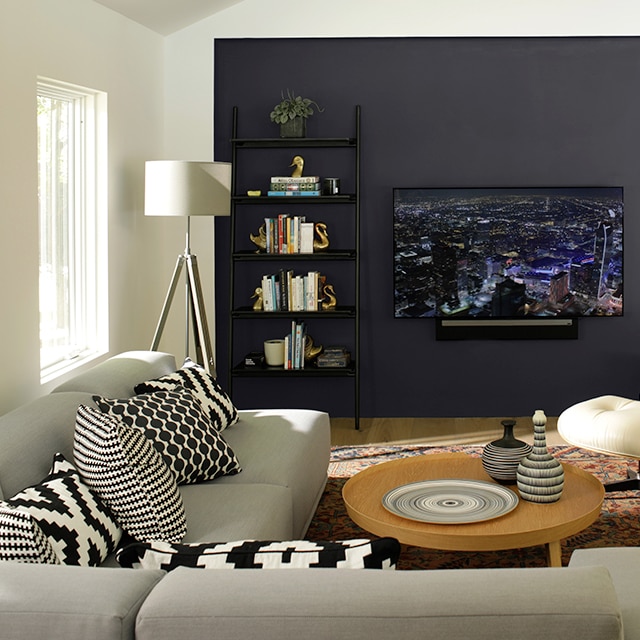A contemporary living room with gray couch, a black-painted accent wall with built-in bookcase, a large screen television, and circular wooden coffee table. A contemporary living room with gray couch, a black-painted accent wall with built-in bookcase, a large screen television, and circular wooden coffee table.
