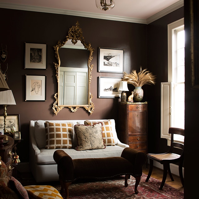 A rich, brown-painted living room with framed artwork and mirror, a loveseat and settee, off-white trim, and a soft pink ceiling. A rich, brown-painted living room with framed artwork and mirror, a loveseat and settee, off-white trim, and a soft pink ceiling.