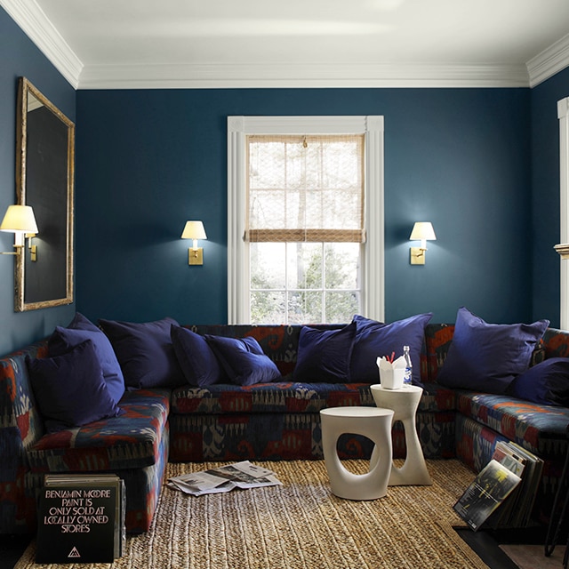 A comfy living room with dark, rich blue painted walls, white ceiling and trim, a multi-colored wrap-around sofa with blue pillows, modern white tables, and a seagrass rug. A comfy living room with dark, rich blue painted walls, white ceiling and trim, a multi-colored wrap-around sofa with blue pillows, modern white tables, and a seagrass rug.