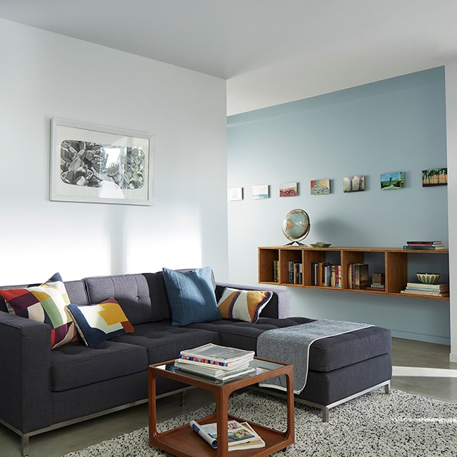 Sun cascading into a light blue and white painted living room with a dark gray sectional sofa and colorful pillows, open to a light blue hallway wall with a mounted bookcase and framed artwork. Sun cascading into a light blue and white painted living room with a dark gray sectional sofa and colorful pillows, open to a light blue hallway wall with a mounted bookcase and framed artwork.