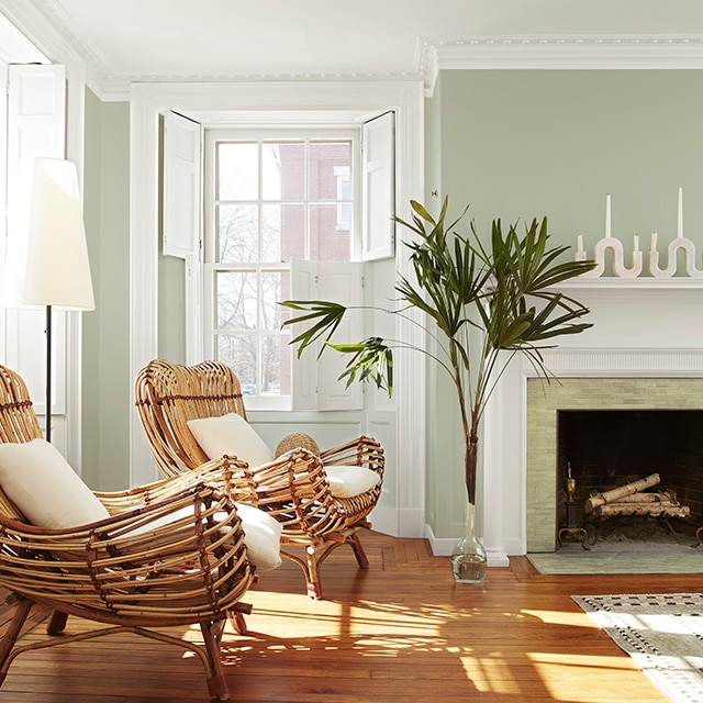 A light green-painted living room wall with beautiful white crown molding, shutters, trim and fireplace mantel, two rattan chairs with white cushions, and tall green palm leaves in a vase. A light green-painted living room wall with beautiful white crown molding, shutters, trim and fireplace mantel, two rattan chairs with white cushions, and tall green palm leaves in a vase.