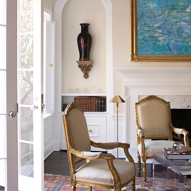 This traditional style living room features neutral tone painted walls with a peach hue, beautiful white decorative molding, open French doors, turquoise framed art over a white fireplace mantel, and two armchairs. This traditional style living room features neutral tone painted walls with a peach hue, beautiful white decorative molding, open French doors, turquoise framed art over a white fireplace mantel, and two armchairs.