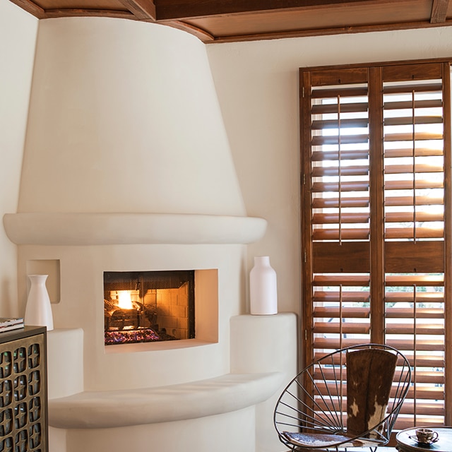 This corner living room space features a beautiful off-white painted adobe style fireplace, off-white walls, a modern metal chair, dark wood plantation shutters, and a dark wood beamed ceiling. This corner living room space features a beautiful off-white painted adobe style fireplace, off-white walls, a modern metal chair, dark wood plantation shutters, and a dark wood beamed ceiling.