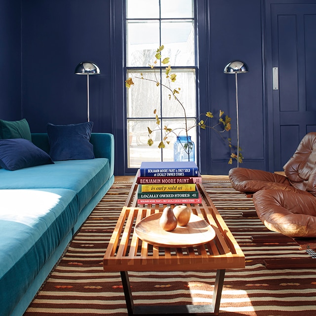 Stunning dark navy blue painted walls and door, and tall center window are backdrop to this inviting living room space with a turquoise sofa, long wood table, and two leather chairs. Stunning dark navy blue painted walls and door, and tall center window are backdrop to this inviting living room space with a turquoise sofa, long wood table, and two leather chairs.